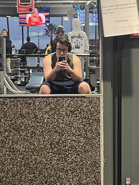 Adrian sitting on a bench taking a mirror selfie at the gym